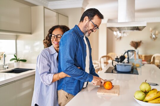 Middle age hispanic couple hugging each other cutting orange at kitchen