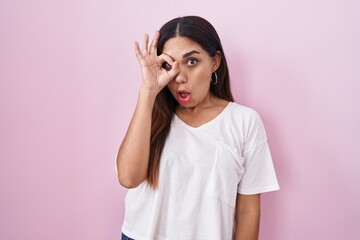 Young arab woman standing over pink background doing ok gesture shocked with surprised face, eye looking through fingers. unbelieving expression.