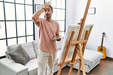 Young hispanic man with beard painting on canvas at home surprised with hand on head for mistake, remember error. forgot, bad memory concept.