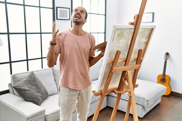 Young hispanic man with beard painting on canvas at home crazy and mad shouting and yelling with aggressive expression and arms raised. frustration concept.