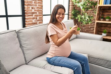 Young latin woman pregnant eating salad sitting on sofa at home