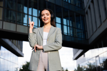 Woman in headphones standing on buisness centre background pointing fingure up speaking with coleaugues listening to music resting after hard day at work.