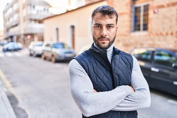 Young hispanic man with serious expression standing with arms crossed gesture at street