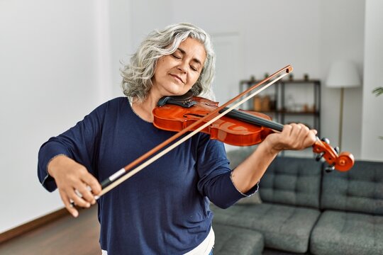 Middle age grey-haired artist woman playing violin standing at home.