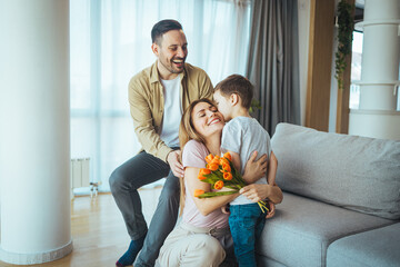 Happy beautiful young mother with flowers and handmade card cuddling adorable small kid son,...