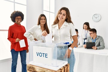 Young voter woman smiling happy putting vote in ballot standing at electoral center.