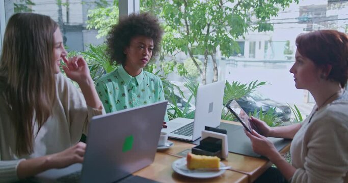 Three Happy Diverse Women Sitting At Coffee Shop Table By Window Overlooking City Sidewalk. Group Of Female Friends Chatting And Laughing In Conversation. Authentic Real Life Laugh And Smile