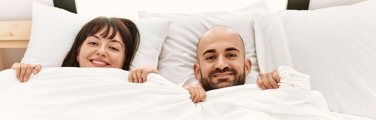 Young hispanic couple covering face with bedsheet lying on the bed at bedroom.