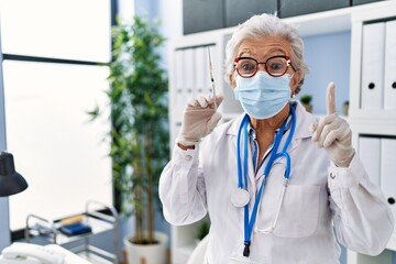 Fototapeta premium Senior woman with grey hair wearing doctor uniform and medical mask holding syringe surprised with an idea or question pointing finger with happy face, number one