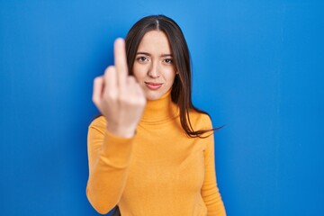 Young brunette woman standing over blue background showing middle finger, impolite and rude fuck...