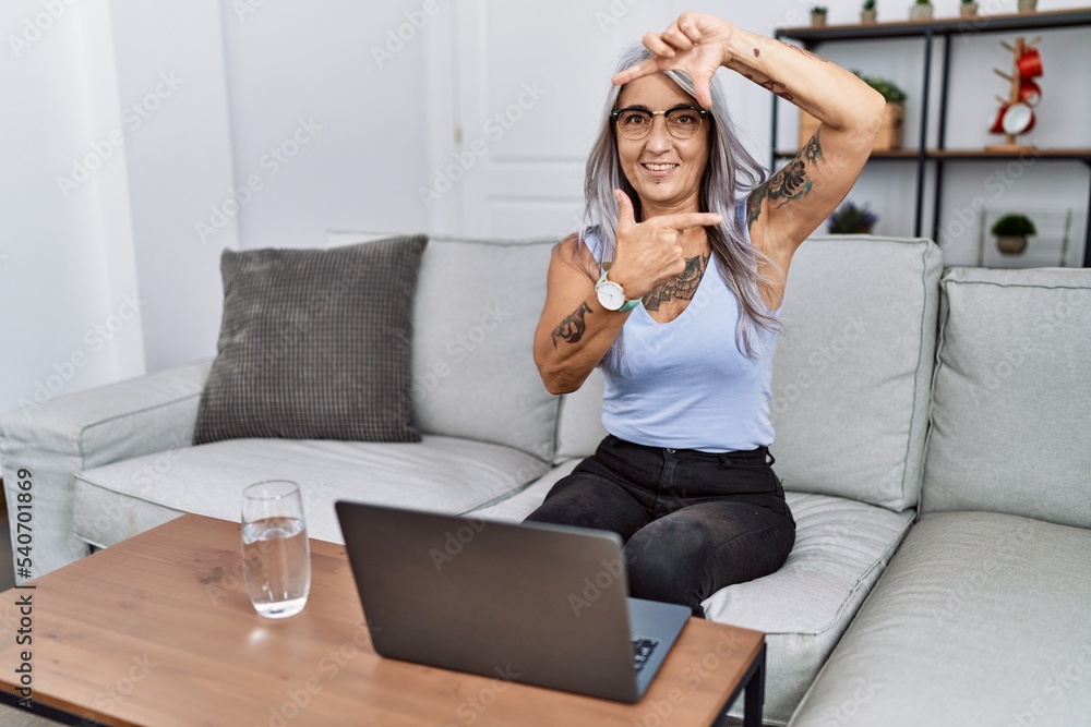 Poster middle age grey-haired woman using laptop at home smiling making frame with hands and fingers with h