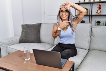 Middle age grey-haired woman using laptop at home smiling making frame with hands and fingers with happy face. creativity and photography concept.