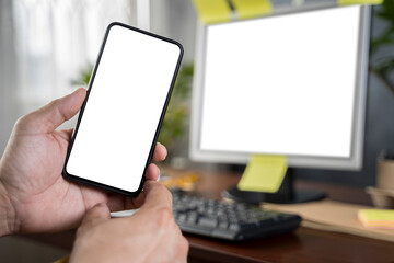 Close up with man's hand holding smartphone in front of laptop, blank screen of smartphone and laptop.