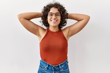 Young hispanic woman wearing glasses standing over isolated background relaxing and stretching, arms and hands behind head and neck smiling happy