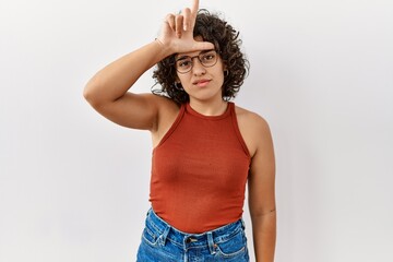 Young hispanic woman wearing glasses standing over isolated background making fun of people with fingers on forehead doing loser gesture mocking and insulting.