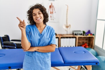 Young hispanic physiotherapist woman working at pain recovery clinic smiling happy pointing with hand and finger to the side