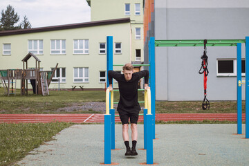 blonde athlete in black sportswear lifts his entire weight using his arms on the bars in an outdoor playground. Strengthening the upper limbs