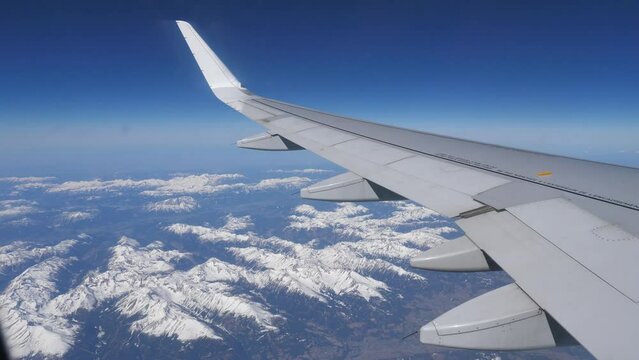 View from passenger cabin window of wing of aircraft flying over Austrian Alps on sunny day against background of blue sky and landscape with snow-capped rocky mountain ranges below. Travel concept