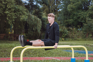 Blonde athlete in black sportswear raises his legs to torso level on the bars to strengthen the abdomen called core. Outdoor gym focused on working with your own body