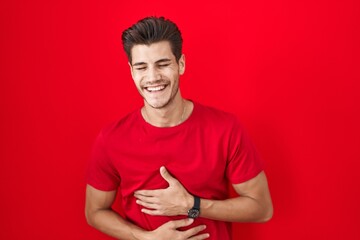 Young hispanic man standing over red background smiling and laughing hard out loud because funny...