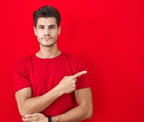 Young hispanic man standing over red background pointing with hand finger to the side showing advertisement, serious and calm face