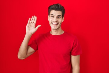 Young hispanic man standing over red background waiving saying hello happy and smiling, friendly welcome gesture