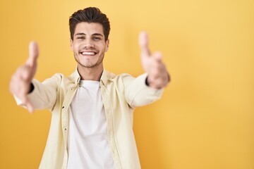 Naklejka premium Young hispanic man standing over yellow background looking at the camera smiling with open arms for hug. cheerful expression embracing happiness.