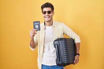 Young hispanic man holding suitcase going on summer vacation holding italian passport smiling and...