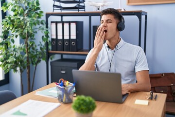Young hispanic man working at the office wearing headphones bored yawning tired covering mouth with hand. restless and sleepiness.