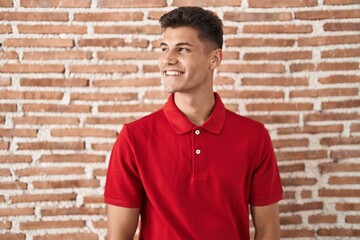 Young hispanic man standing over bricks wall smiling looking to the side and staring away thinking.