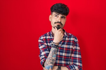Young hispanic man with beard standing over red background with hand on chin thinking about...