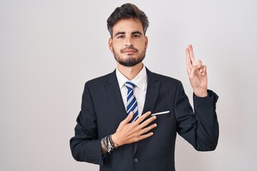 Young hispanic man with tattoos wearing business suit and tie smiling swearing with hand on chest and fingers up, making a loyalty promise oath