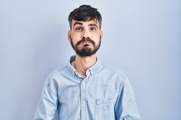 Young hispanic man with beard standing over blue background puffing cheeks with funny face. mouth inflated with air, crazy expression.