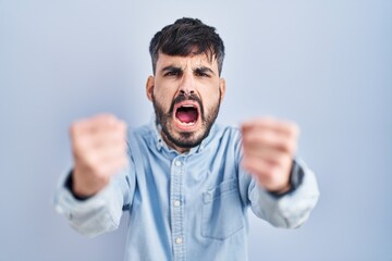 Young hispanic man with beard standing over blue background angry and mad raising fists frustrated and furious while shouting with anger. rage and aggressive concept.