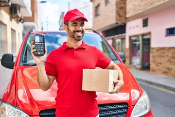 Young hispanic man with beard wearing delivery uniform and cap holding dataphone winking looking at...