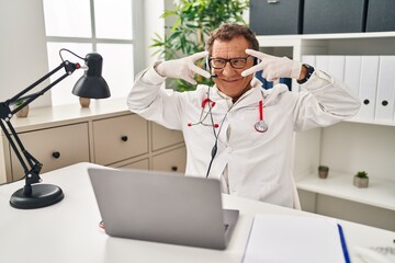 Naklejka na ściany i meble Senior doctor man working on online appointment doing peace symbol with fingers over face, smiling cheerful showing victory