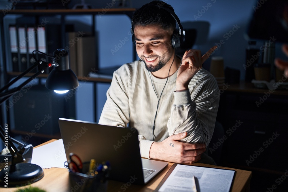 Sticker Young handsome man working using computer laptop at night with a big smile on face, pointing with hand and finger to the side looking at the camera.