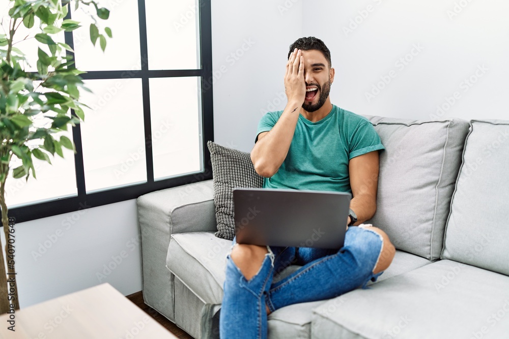 Wall mural young handsome man with beard using computer laptop sitting on the sofa at home covering one eye wit