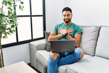 Young handsome man with beard using computer laptop sitting on the sofa at home smiling with hands on chest with closed eyes and grateful gesture on face. health concept.