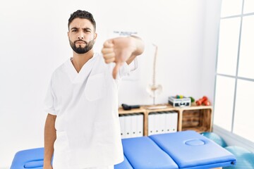 Young handsome man with beard working at pain recovery clinic looking unhappy and angry showing rejection and negative with thumbs down gesture. bad expression.