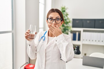 Young brunette doctor woman holding glass of water covering mouth with hand, shocked and afraid for mistake. surprised expression