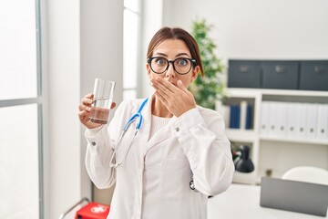 Young brunette doctor woman holding glass of water covering mouth with hand, shocked and afraid for...