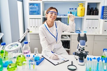 Young hispanic woman wearing scientist uniform holding test tube and writing on clipboard at laboratory