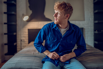 A young man in dark-blue shirt sitting on the bed - obrazy, fototapety, plakaty