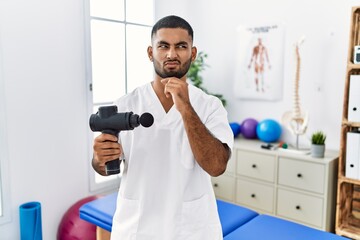 Fototapeta na wymiar Young indian physiotherapist holding therapy massage gun at wellness center thinking worried about a question, concerned and nervous with hand on chin
