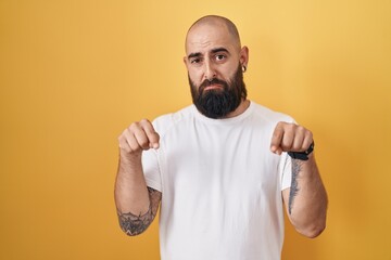 Young hispanic man with beard and tattoos standing over yellow background pointing down looking sad and upset, indicating direction with fingers, unhappy and depressed.