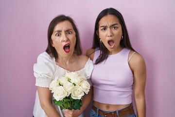 Hispanic mother and daughter holding bouquet of white flowers in shock face, looking skeptical and sarcastic, surprised with open mouth
