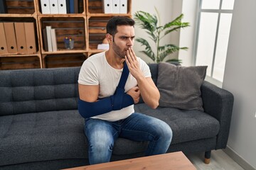 Young hispanic man with beard wearing arm on sling sitting at therapy consult covering mouth with...