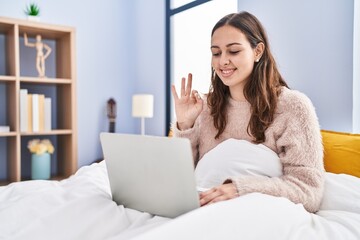 Young hispanic woman using computer laptop on the bed doing ok sign with fingers, smiling friendly gesturing excellent symbol