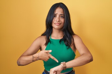 Brunette woman standing over yellow background in hurry pointing to watch time, impatience, upset and angry for deadline delay